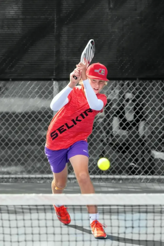 young boy wearing red pickleball shirt and cap in mid-swing playing pickleball