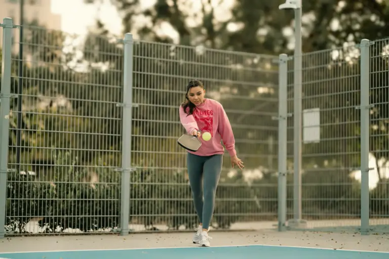 young woman with long brown hair wearing a pink sweater and dark leggins playing pickleball in mid-swing