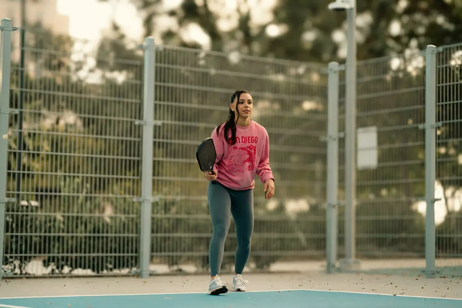 young woman with long brown hair wearing a pink sweater and dark leggins playing pickleball